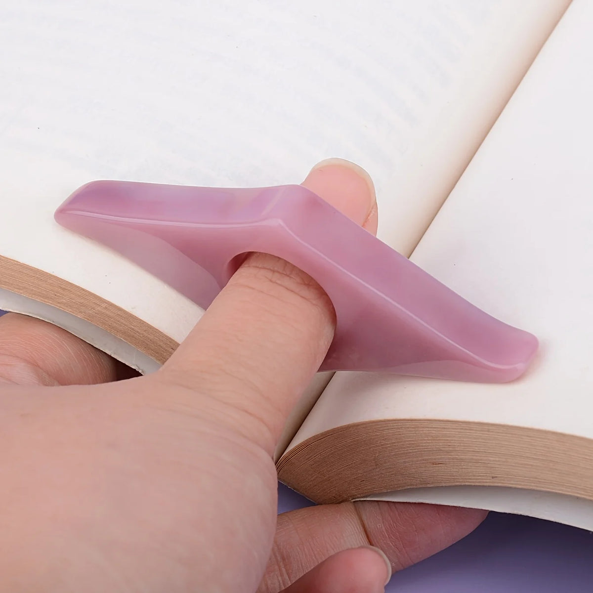 A hand holds a translucent purple resin thumb book stand resting on a white page.