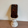 A brown patterned phone case for an iPhone is displayed in a white ceramic pedestal.