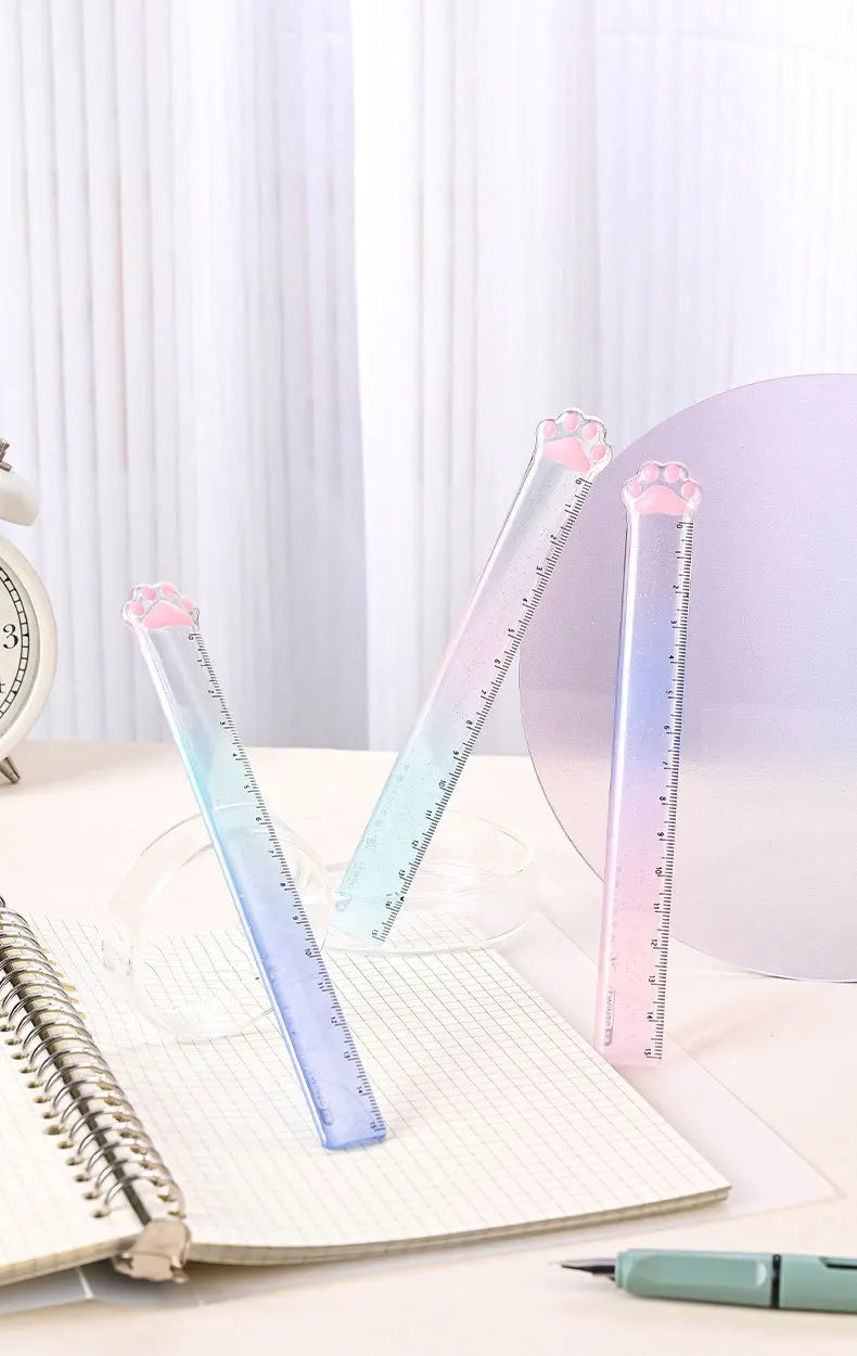 A set of three clear glass rulers shaped like cat paws with a gradient color, resting on an open notebook.