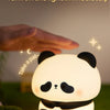 A white panda silicone night light is shown glowing on green grass, with a hand gently petting it.