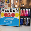 A box of a 24-color gouache paint set with tubes and a wooden shelf in the background.