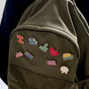 A close-up of a dark olive green fabric featuring several enamel pins with text like NO MORE DRAMA and 80s Baby.