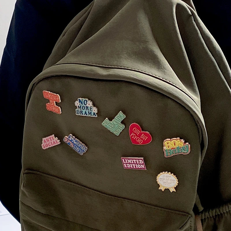 A close-up of a dark olive green fabric featuring several enamel pins with text like NO MORE DRAMA and 80s Baby.