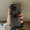 A hand holds a dark gray phone case featuring a black spider web design and a silver watch.