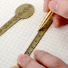 A hand holds a vintage brass straight ruler with a minimalist design placed on a graph paper background.