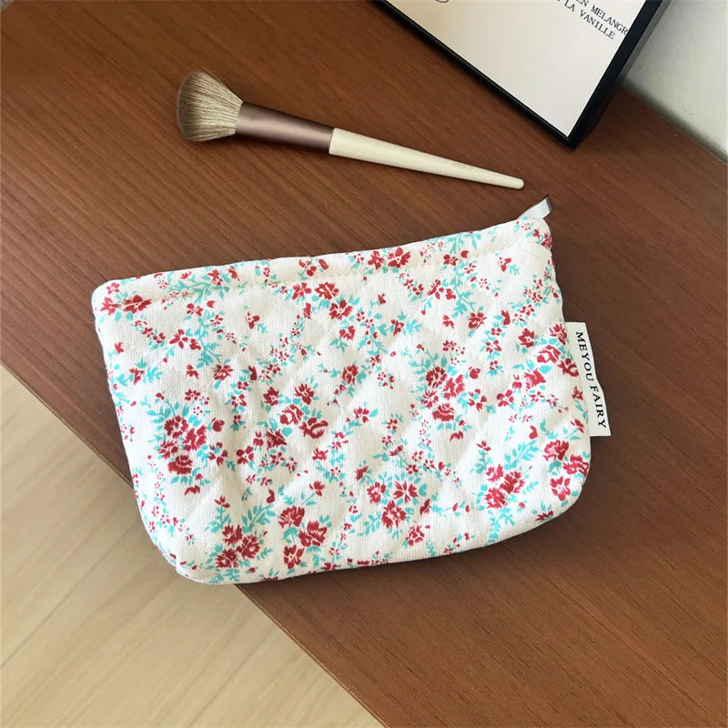 A floral quilted ladies clutch purse with red and blue flowers rests on a wooden surface next to a makeup brush.