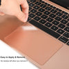 A hand applies a thin, rose gold trackpad protector to the surface of a MacBook laptop.