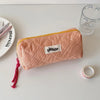 A pink checkered pencil case with a yellow trim is shown next to a glass and a silver ring on a white surface.