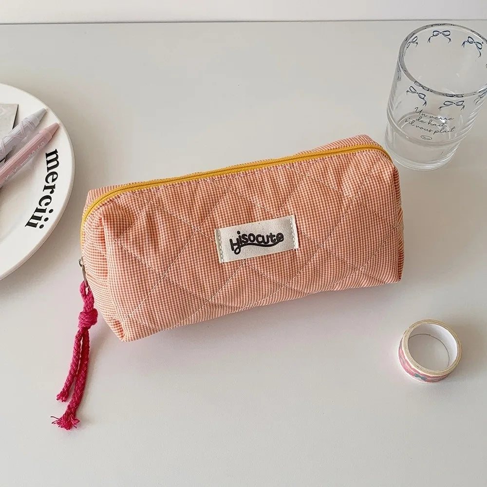A pink checkered pencil case with a yellow trim is shown next to a glass and a silver ring on a white surface.