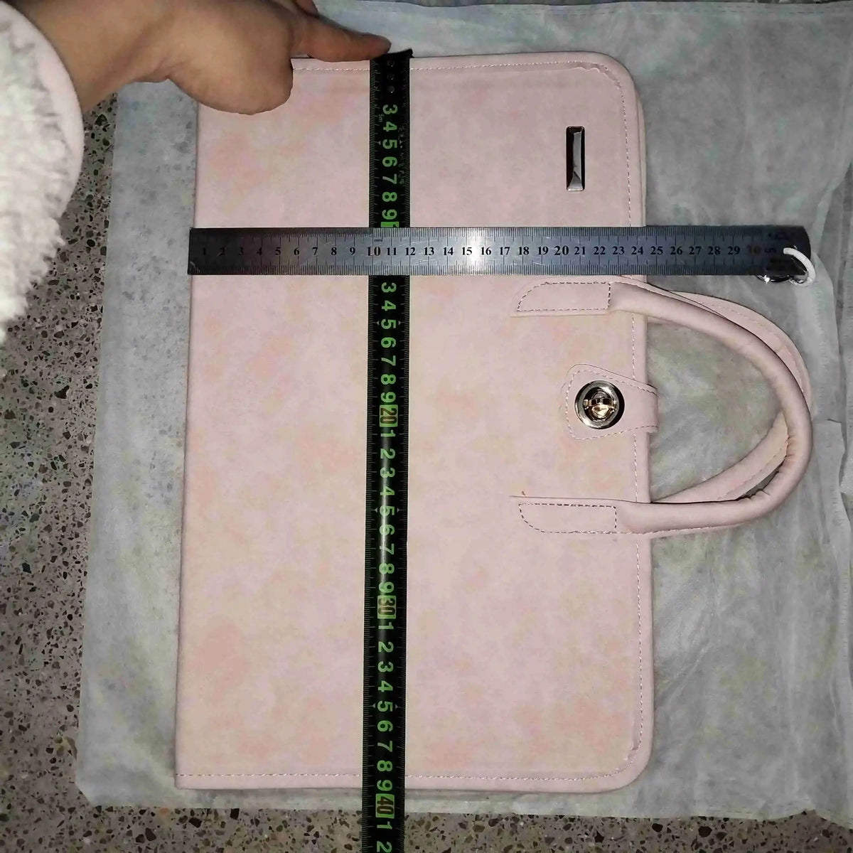 A pink laptop sleeve with a handle is shown lying on a gray fabric surface, measured by a ruler.