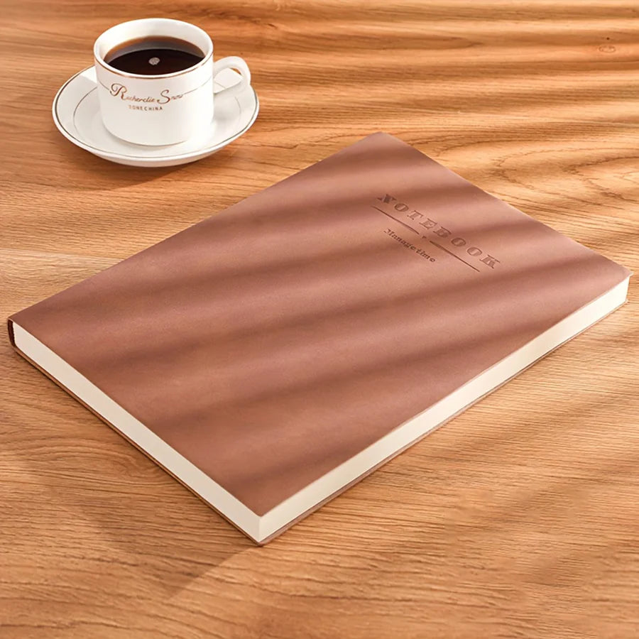 A brown notebook lies on a wooden surface next to a white cup and saucer.