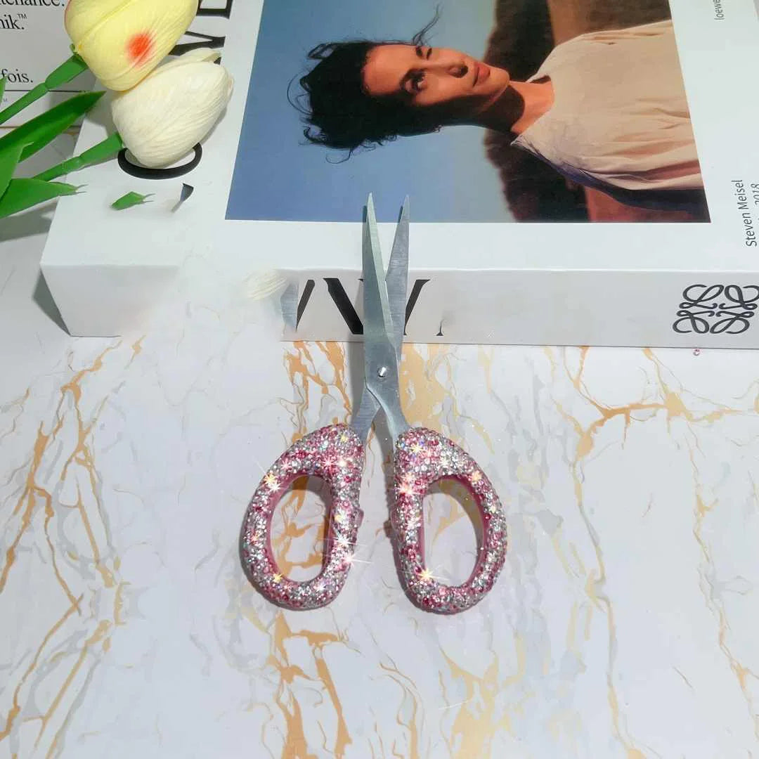 A pair of pink rhinestone crafting scissors resting on a white surface next to a box and a yellow flower.