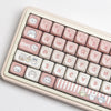 A close-up view of a light pink mechanical keyboard featuring white and pastel themed dye-sublimated PBT keycaps.