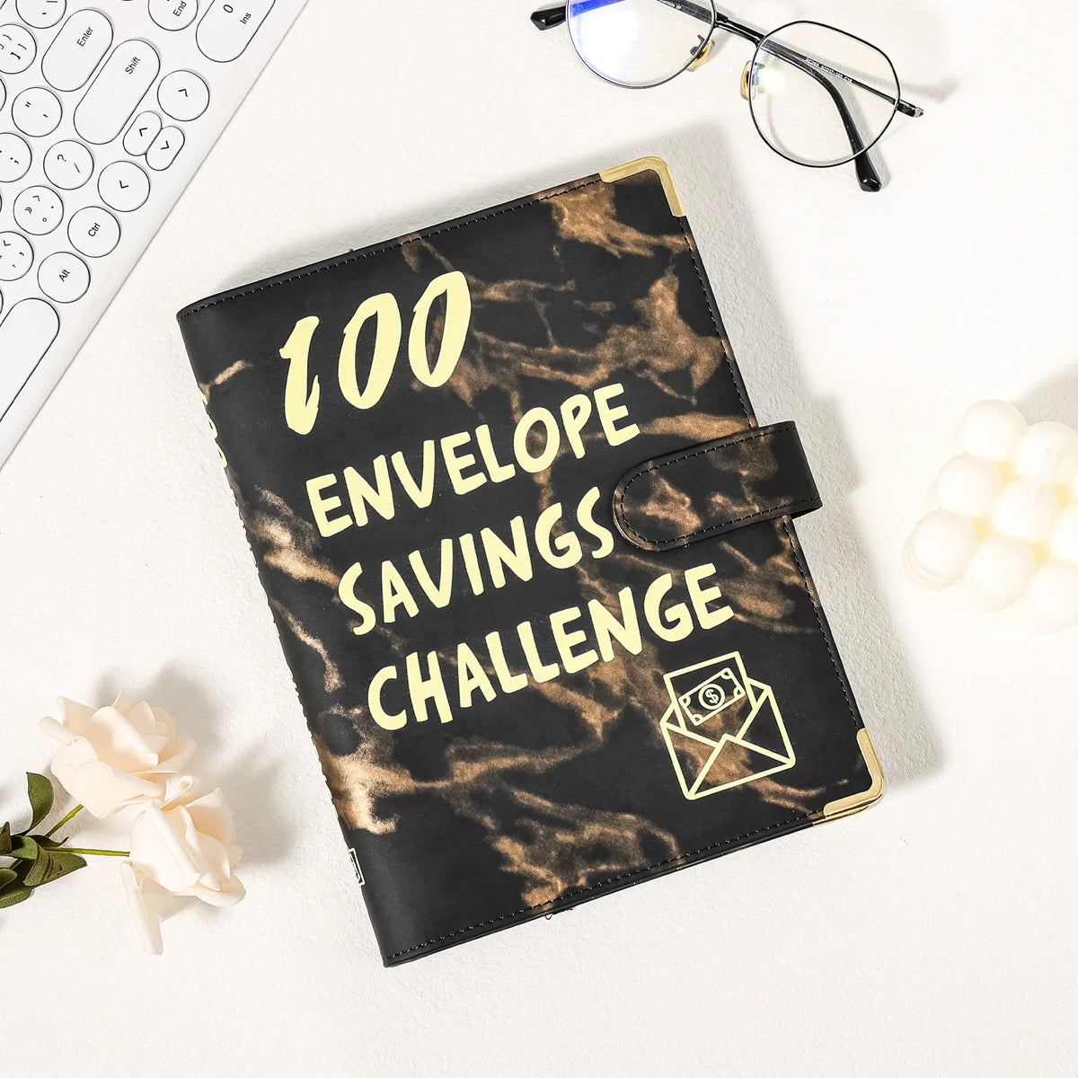 A black marble budget binder titled 100 Envelope Savings Challenge resting on a white surface.