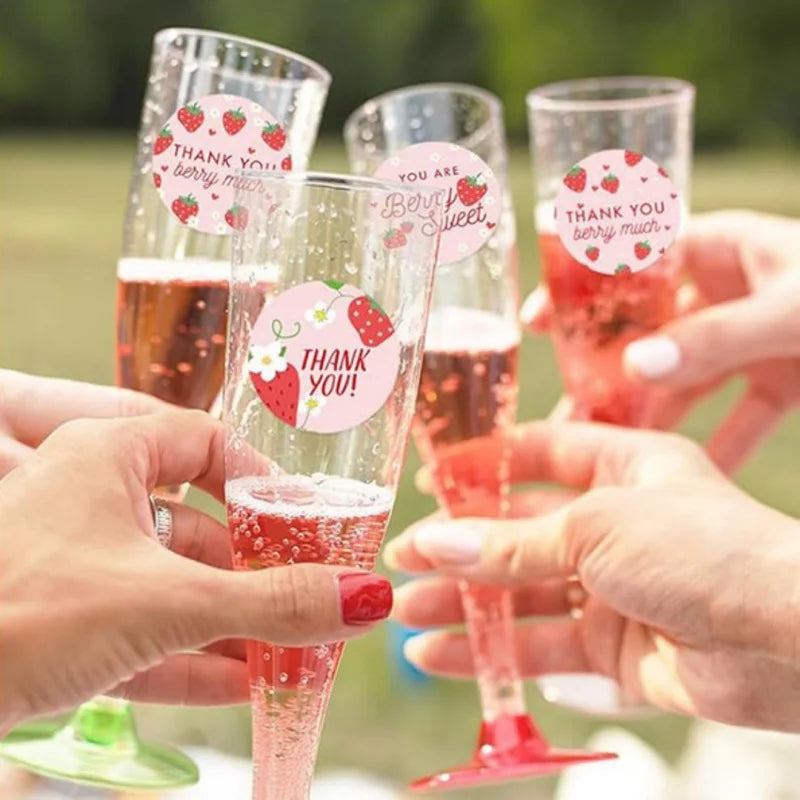 Hands hold stemmed glasses filled with red liquid and strawberry adhesive stickers