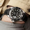 A close-up of a black watch with a silver bezel and leather band worn on a wrist, visible under a beige jacket sleeve.