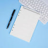 Un refil de cuaderno de hojas sueltas blanco junto a un bolígrafo negro y un teclado de portátil plateado sobre una superfici