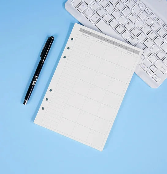 Un refil de cuaderno de hojas sueltas blanco junto a un bolígrafo negro y un teclado de portátil plateado sobre una superfici