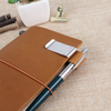 A brown leather journal is clipped shut with a brass pen holder and clip on a beige fabric surface.