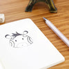A white A6 blank sketchbook with a simple black line drawing of a horned animal and a silver pen on a wooden surface.