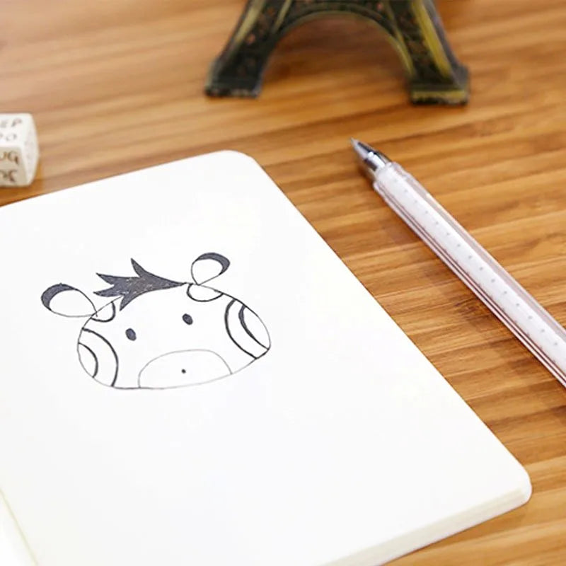 A white A6 blank sketchbook with a simple black line drawing of a horned animal and a silver pen on a wooden surface.