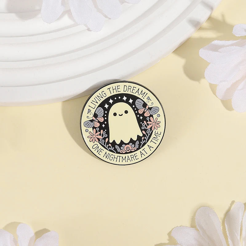 A round enamel pin featuring a cute ghost character surrounded by flowers and text on a light yellow background.