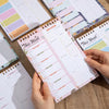 A hand holds a pink weekly meal planner and grocery list pad with organized meal plans and notes.