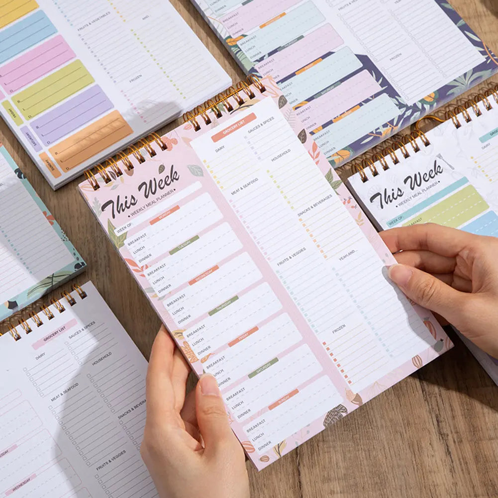 A hand holds a pink weekly meal planner and grocery list pad with organized meal plans and notes.