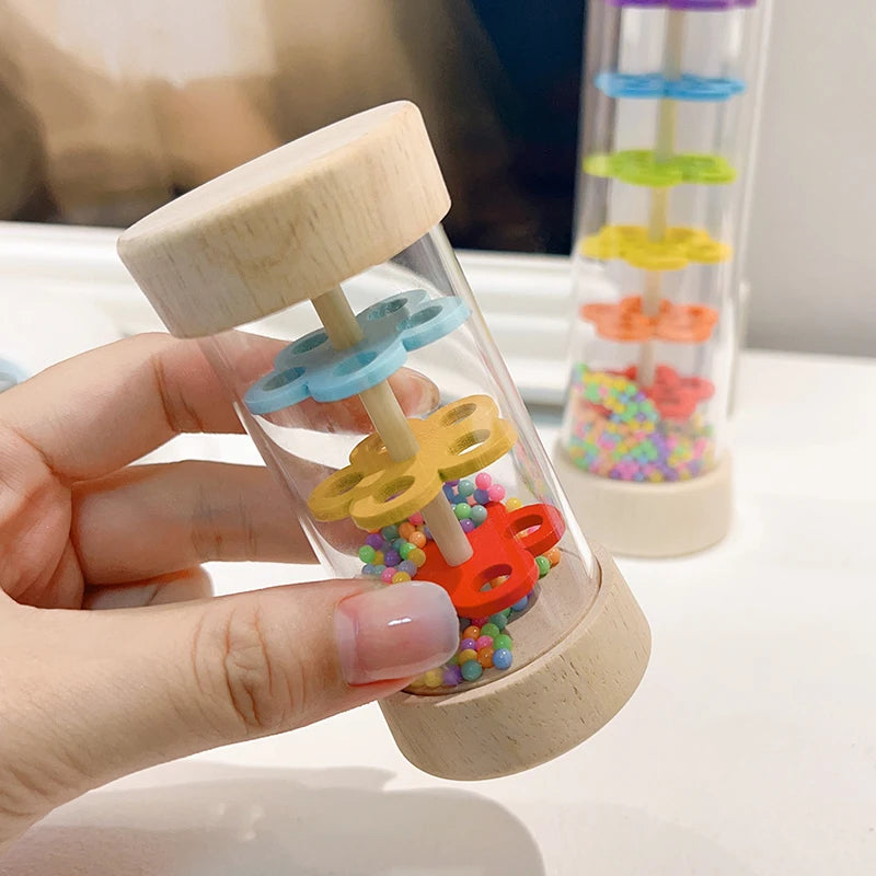 A hand holds a wooden rain sound tube musical instrument filled with colorful beads, demonstrating its layered sound mechanis