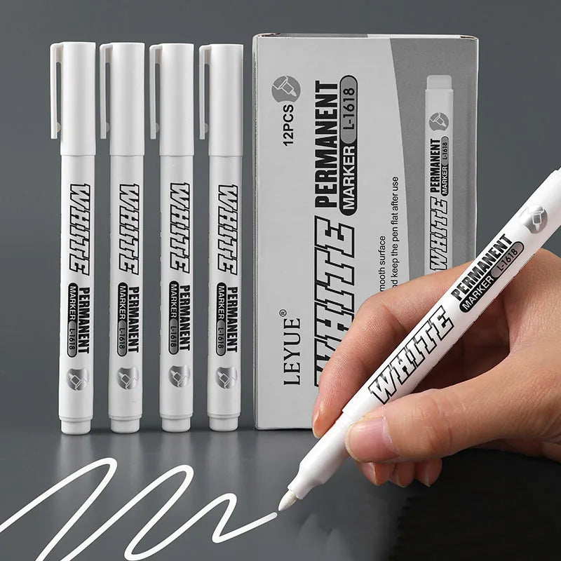 A hand holds a white permanent marker pen next to a box containing twelve permanent markers.