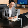 A man is using an adjustable aluminum tablet stand to write in a notebook at a desk.