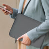 A person in a light gray blazer holds a gray protective sleeve briefcase and a smartphone.