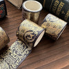 A set of metallic gold washi tape rolls with intricate patterns lying on a wooden surface next to a book.