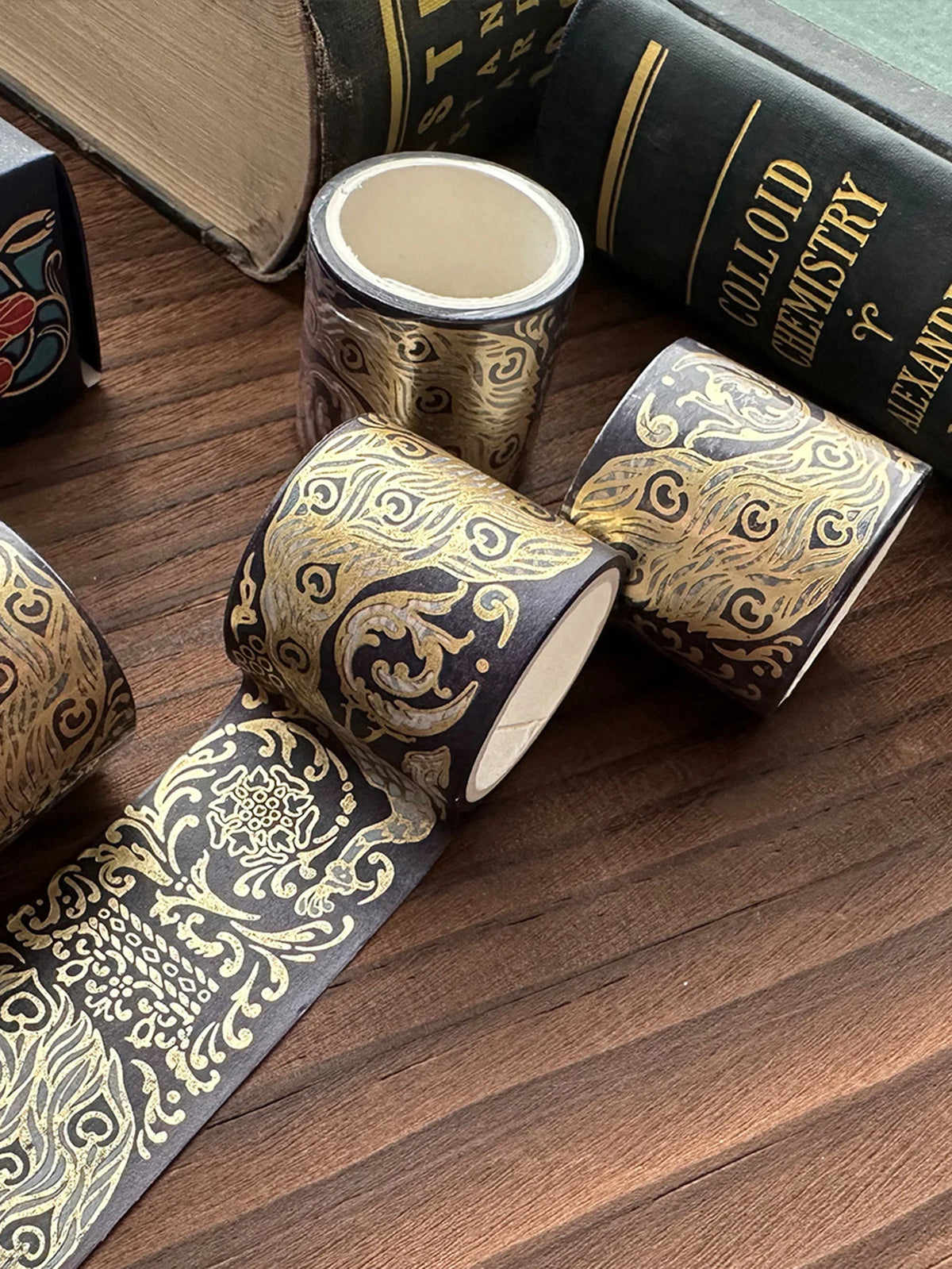 A set of metallic gold washi tape rolls with intricate patterns lying on a wooden surface next to a book.