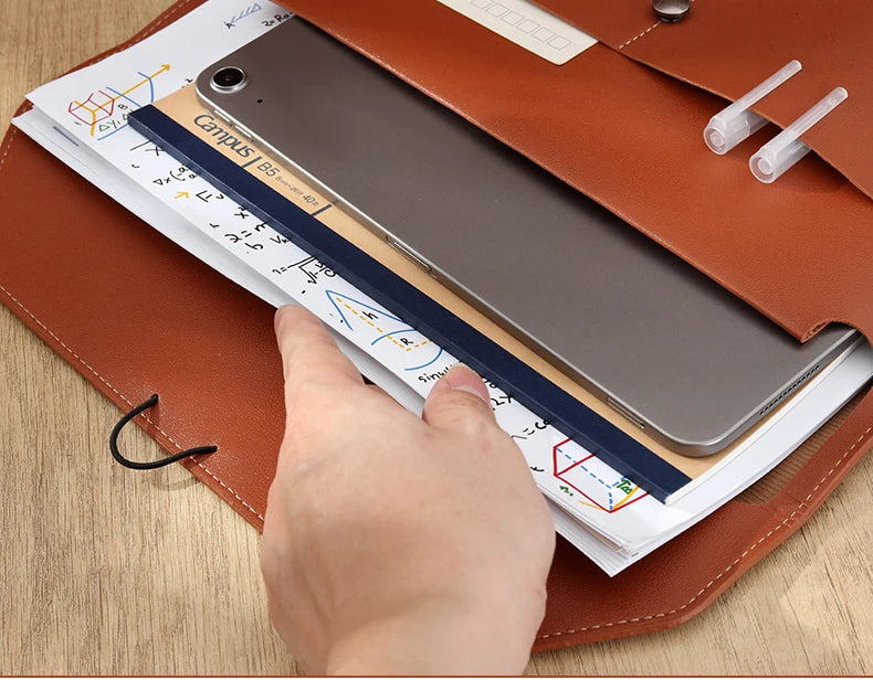 A hand is placing a silver tablet into a leather document organizer bag alongside A4 files.