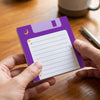 A purple sticky note pad with a cosmic moon theme and a silver pen is held in a person's hand on a wooden table.