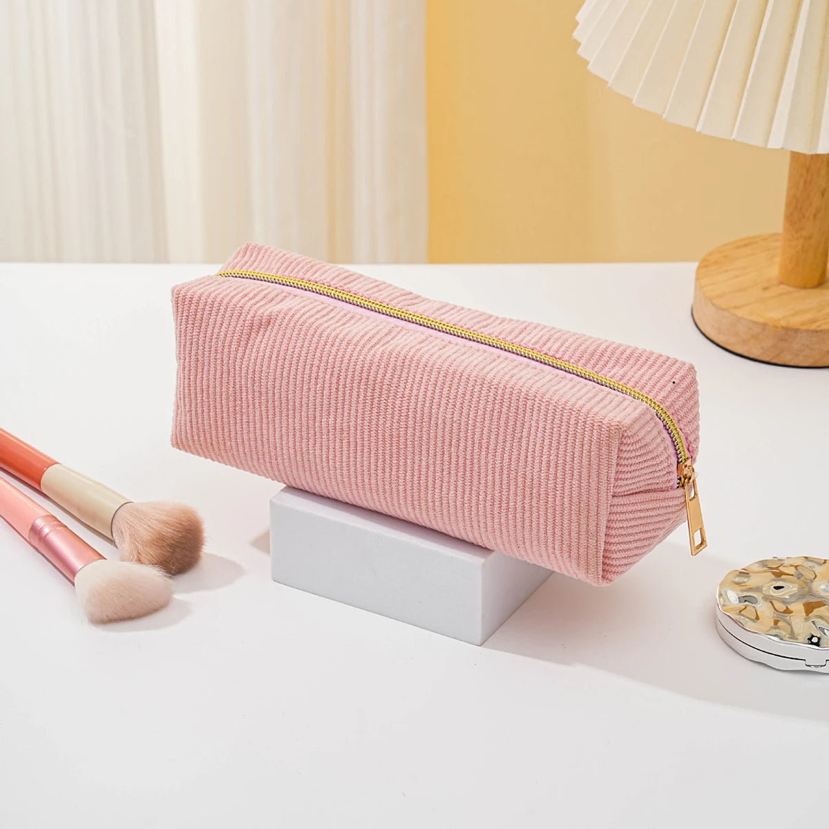 A pink textured pencil case with a gold zipper rests on a white surface next to makeup brushes.