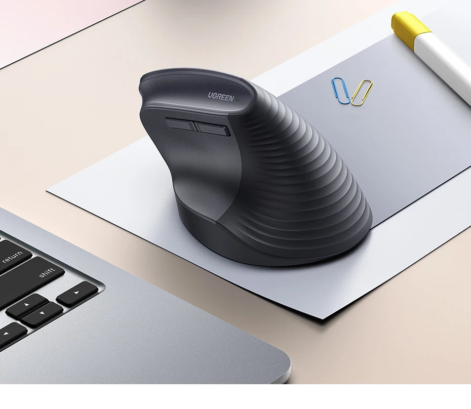A black ergonomic vertical wireless mouse is shown resting on a white sheet of paper next to a silver laptop.