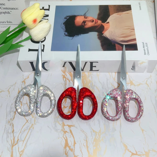 A set of luxury rhinestone crafting scissors with red and pink glitter handles resting on a white box.
