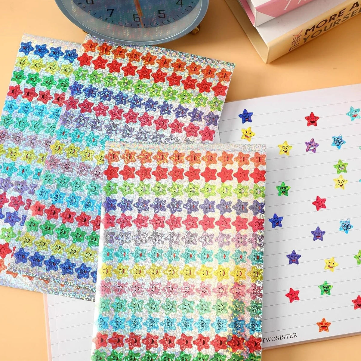 A sheet of colorful glitter foil star stickers for crafts and rewards is shown next to a spiral notebook.