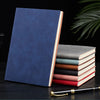 A stack of premium leather journals in various shades of blue, gray, and pink resting on a dark surface with a gold pen.