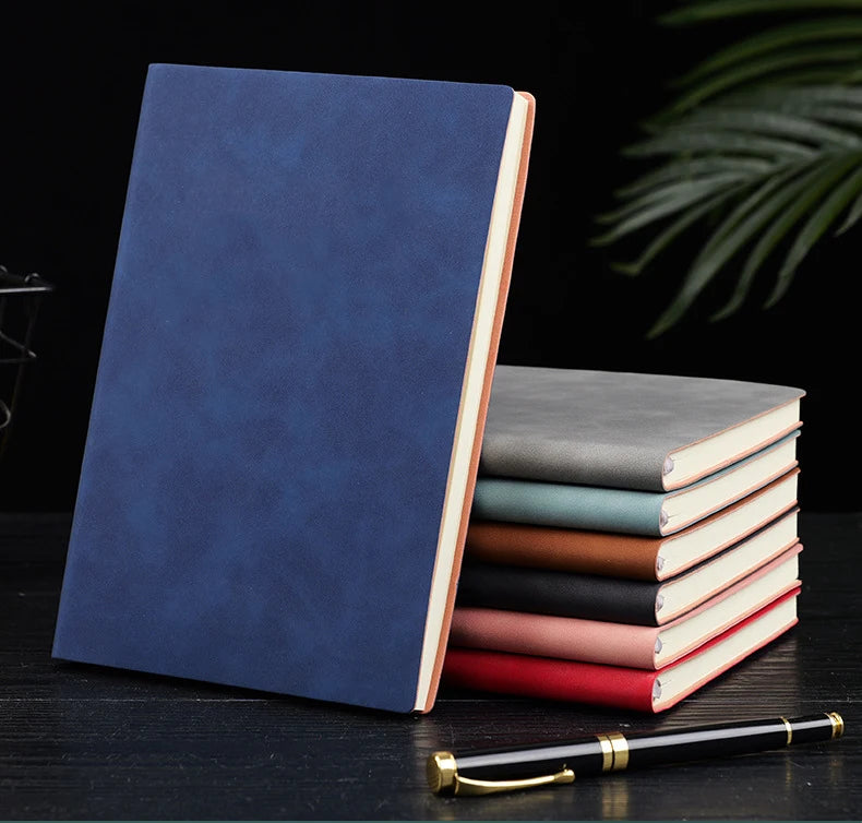 A stack of premium leather journals in various shades of blue, gray, and pink resting on a dark surface with a gold pen.