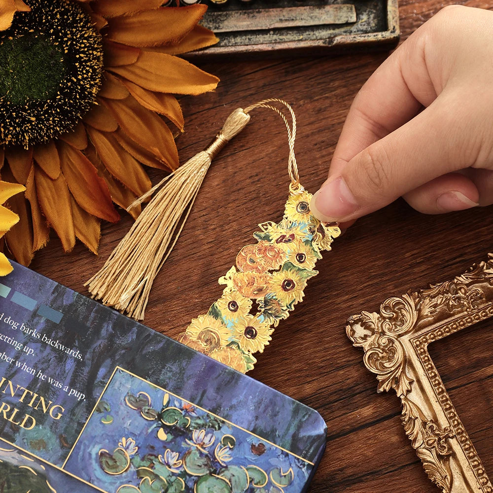 A vintage artistic metal bookmark with a gold tassel resting on a wooden surface next to a sunflower.