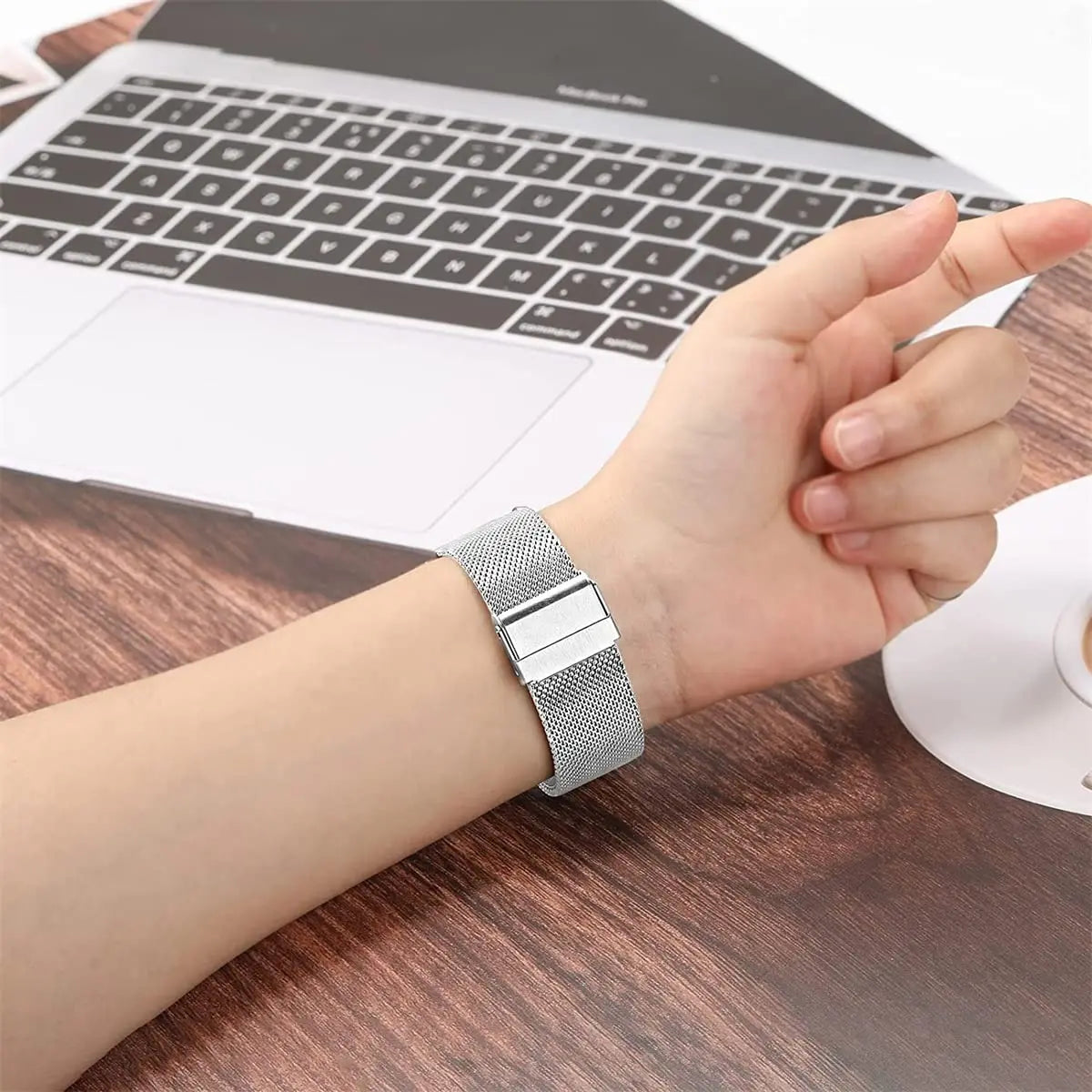 A close-up of a stainless steel Milanese loop watch band on a wrist next to a laptop.