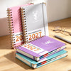 A stack of spiral-bound weekly and monthly planners in pink, gray, and purple with a deer design on a wooden surface.