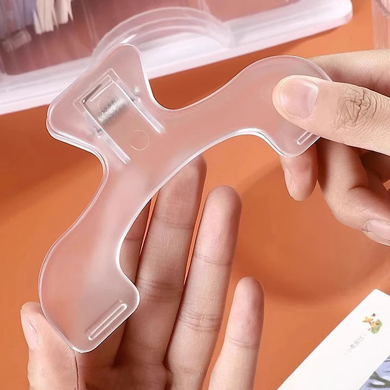 A hand holds a clear, plastic book page clip designed in the shape of a stylized animal or hook.