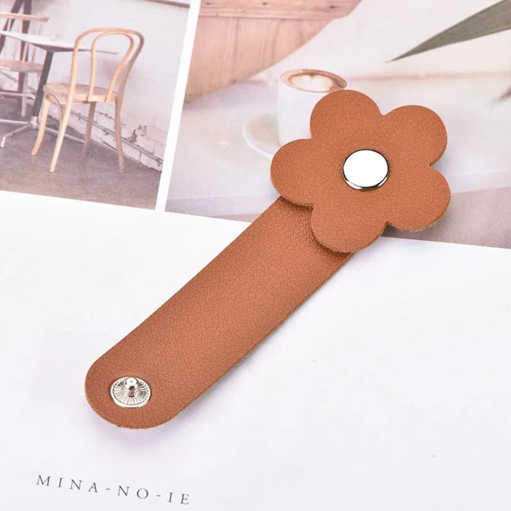 A brown leather cable organizer shaped like a flower with a silver accent, shown next to a wooden chair.