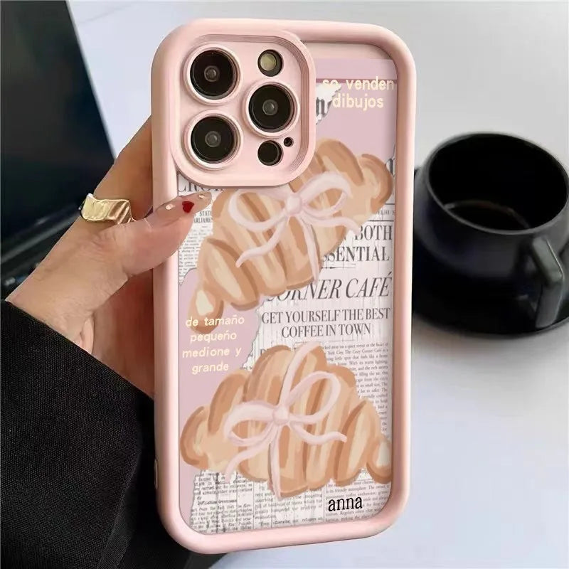 A pink phone case with a croissant newspaper pattern is held by a hand next to a black mug.