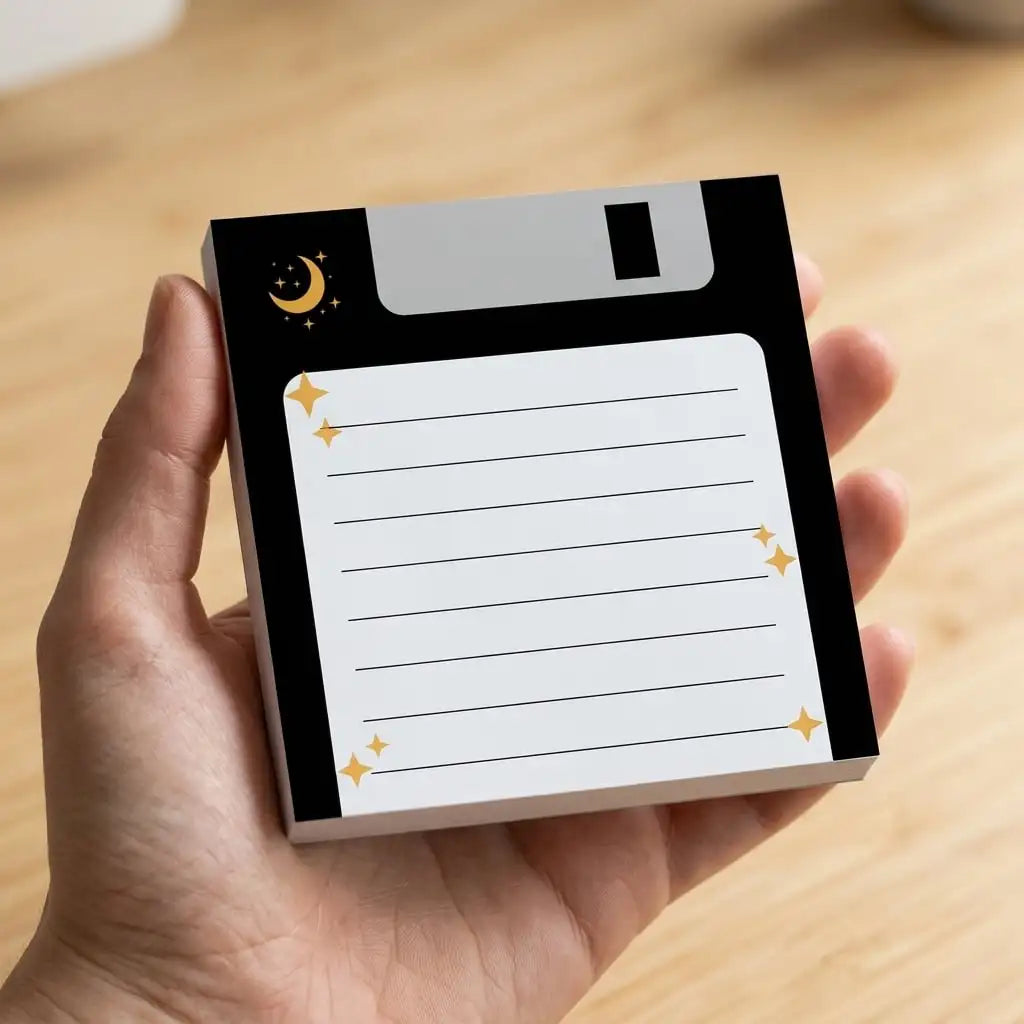 A hand holds a pack of retro floppy disk sticky notes with a cosmic moon theme and a black and white design.