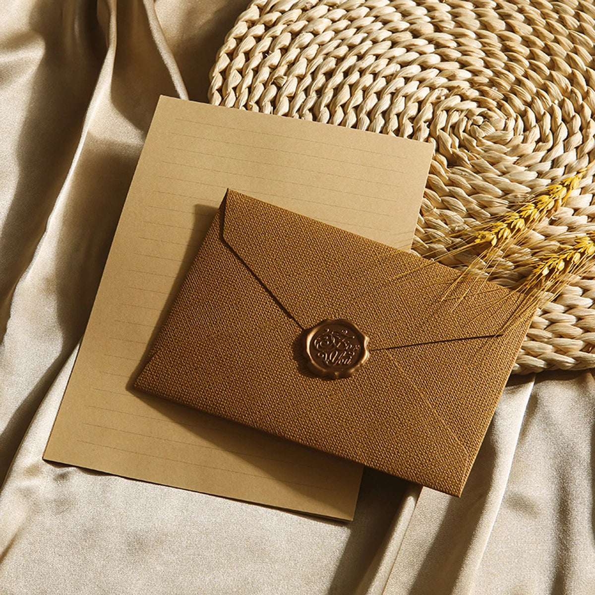 Ein Set brauner Kraftpapierkuvert und ein Brief mit Wachssiegel auf beige Stoff neben einer geflochtenen Strohmatte.